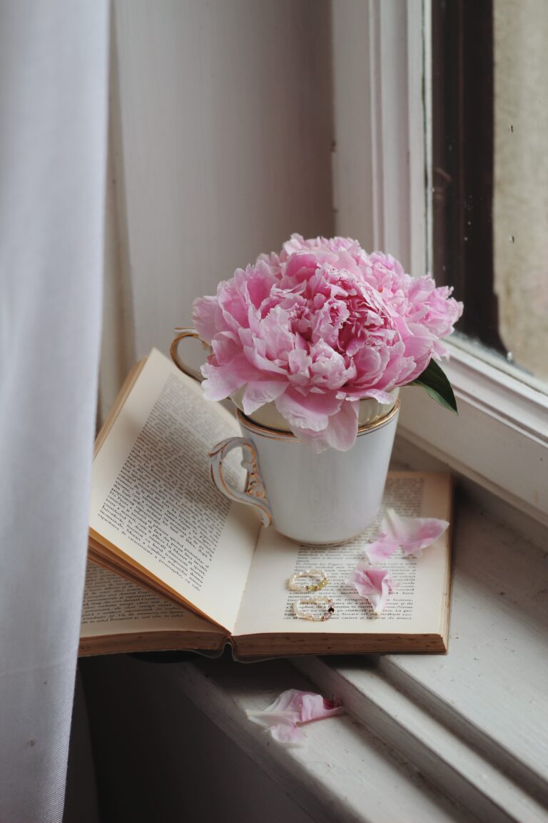 Picture featuring a white tea cup filled with pink peonies sitting atop an open book